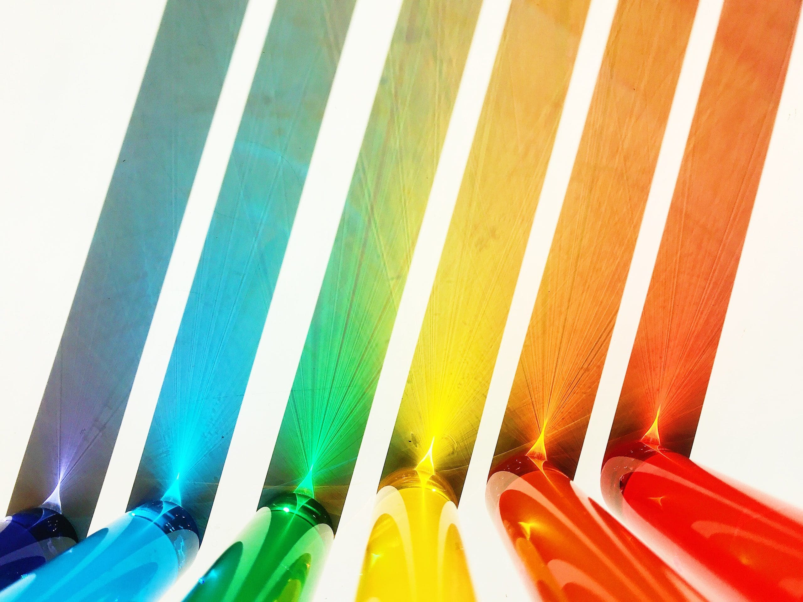 From left to right - 6 coloured semi translucent rods being lit from behind, the rods are navy, light blue, green, yellow, orange and red and the effect of being lit from behind is creating colourful shadows.